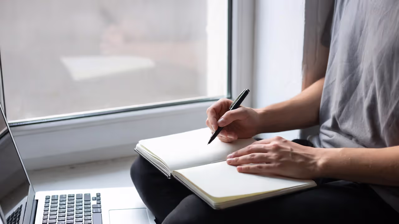 Professional journaling by a window with a closed laptop nearby, symbolizing reflection and redefining success