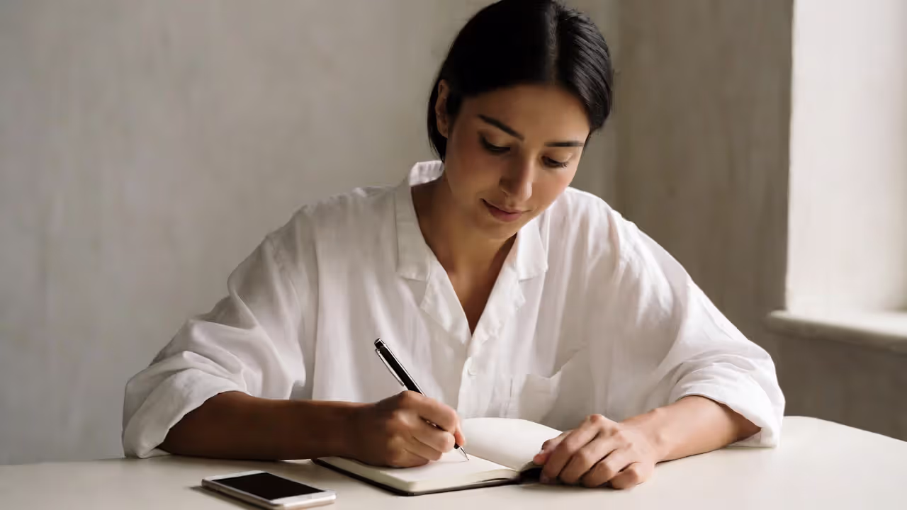 Person journaling at a minimalist desk with a phone face down, calm and reflective.
